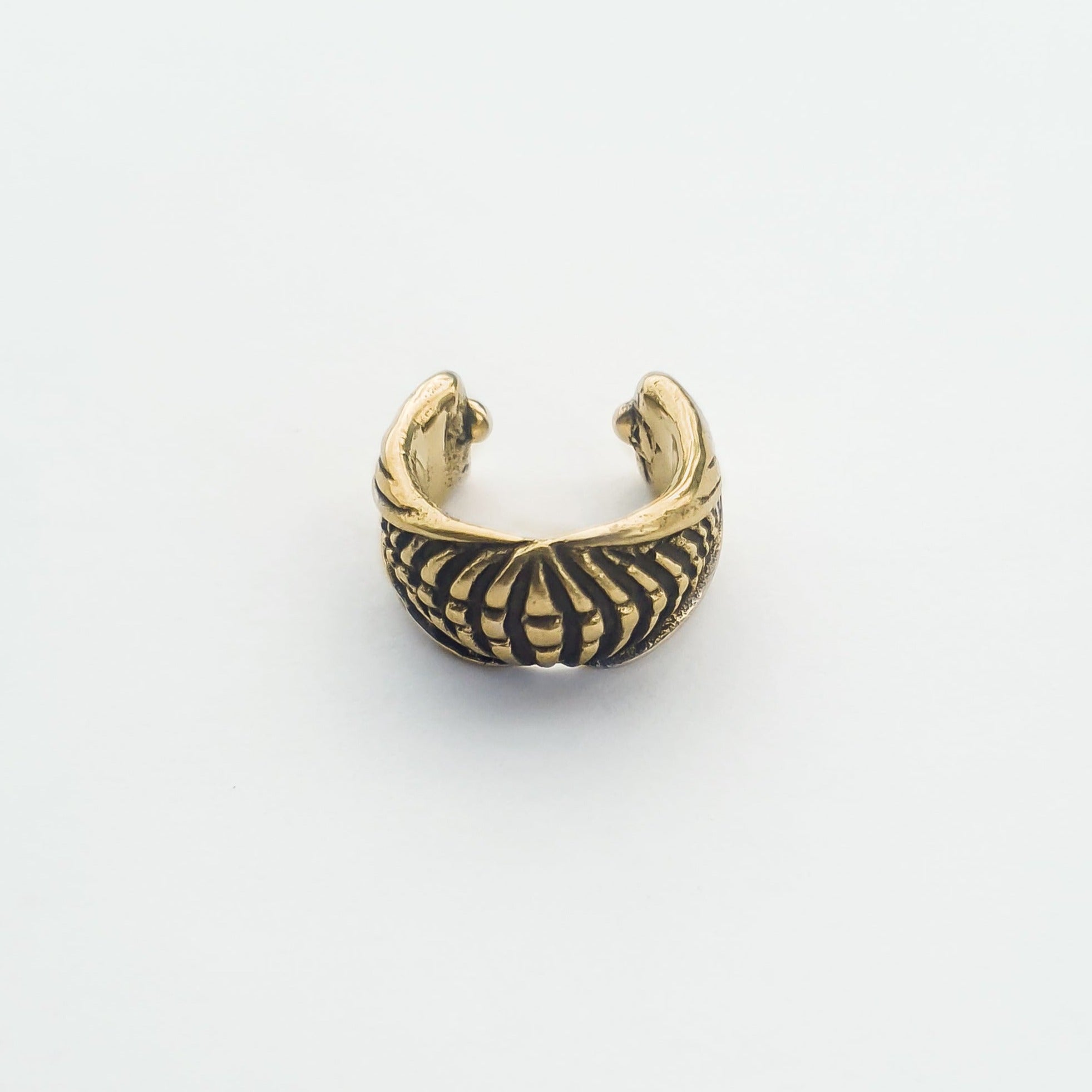 A close-up view of the DARMA Fake Septum Ring or Ear Cuff in Gold by Kaya Movement. This intricately designed, open-ended piece features a series of evenly spaced vertical lines and an elaborate pattern, giving it a vintage or antique appearance. This versatile unisex jewelry is set against a plain, light background.